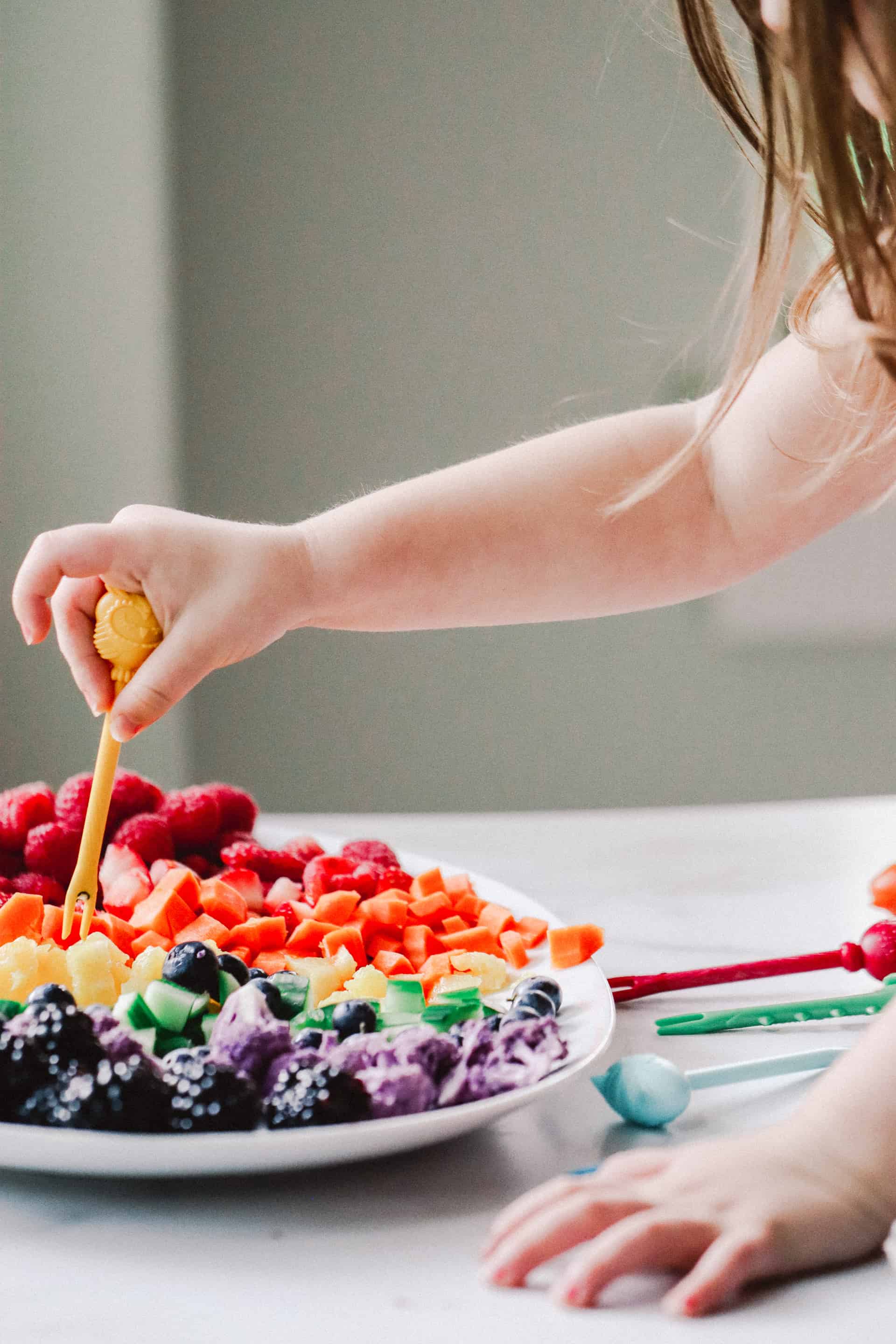 toddler using food pick to eat fruit and vegetables on white platter