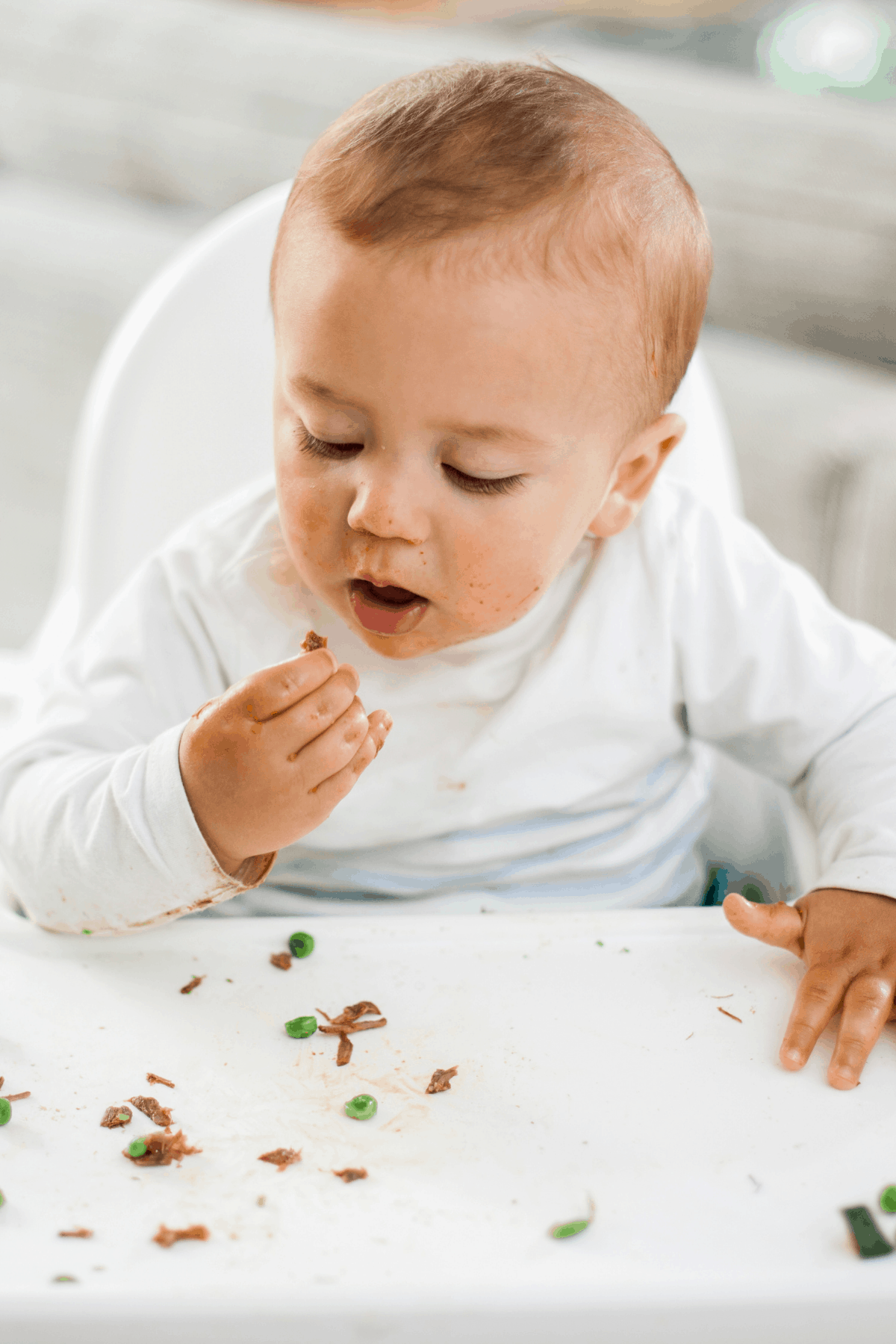 9 month old baby eating finger foods peas and shredded beef in highchair