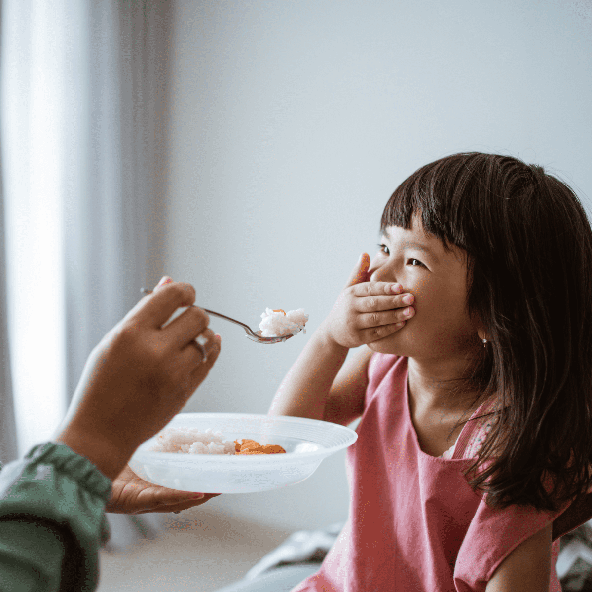 Toddler girl covering her mouth with hand, refusing food from caregiver's hand trying to feed her.