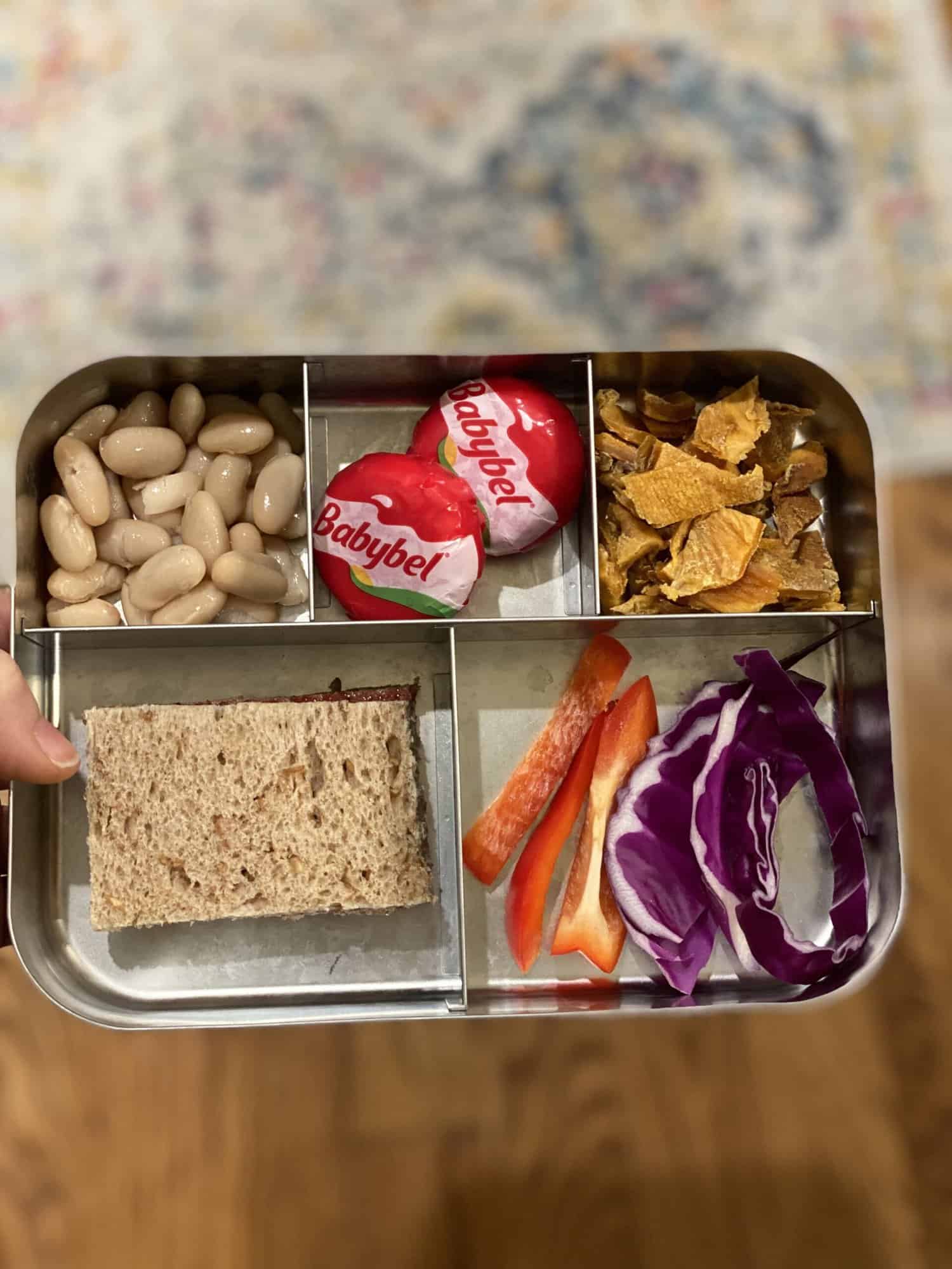 packed lunch for 2 year old with a sandwich on whole wheat bread, babybel cheese, white beans, dried mango, red pepper and cabbage