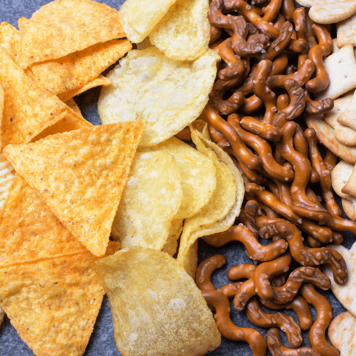 Close-up photo of triangular chips, potato chips, wavy pretzels and small assorted crackers on a dark background. 