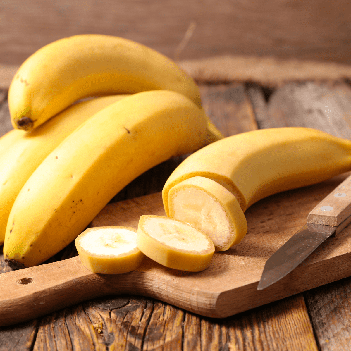 Wooden tabletop with three whole bananas next to a wooden cutting board and wooden-handled knife with a fourth banana being cut into slices with the peel on.