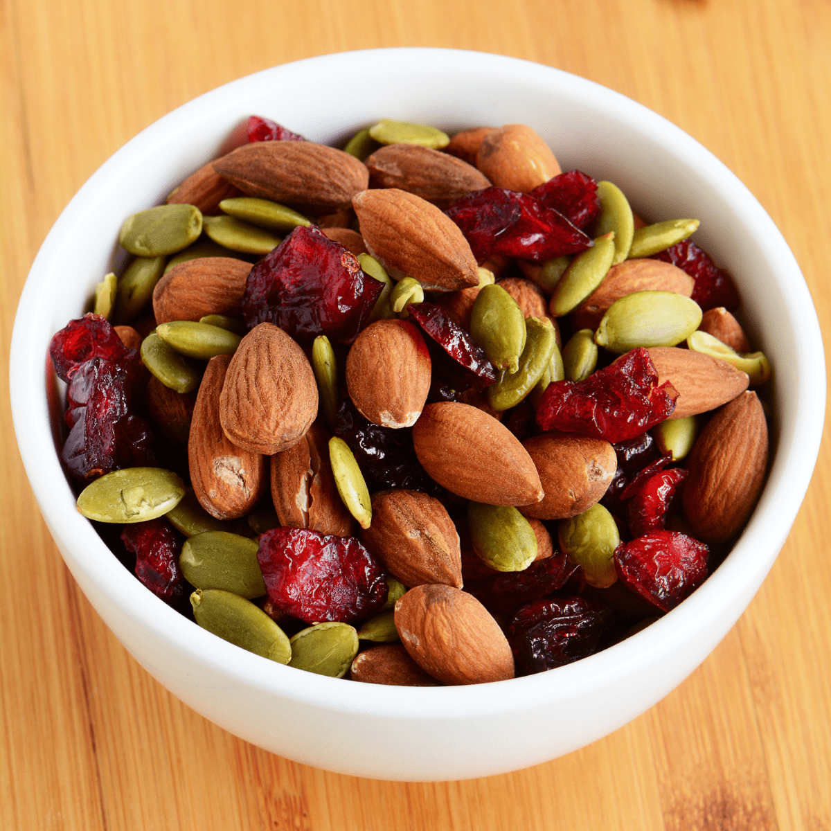 White bowl filled with almonds, seeds and dried cranberries on a wooden table top. 