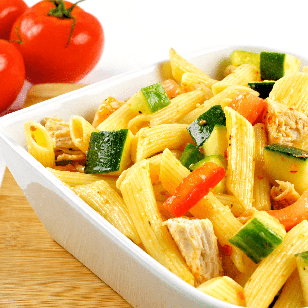 Square white bowl filled with rigatoni noodles, diced chicken and mixed vegetables on wooden tabletop with whole tomatoes in the background