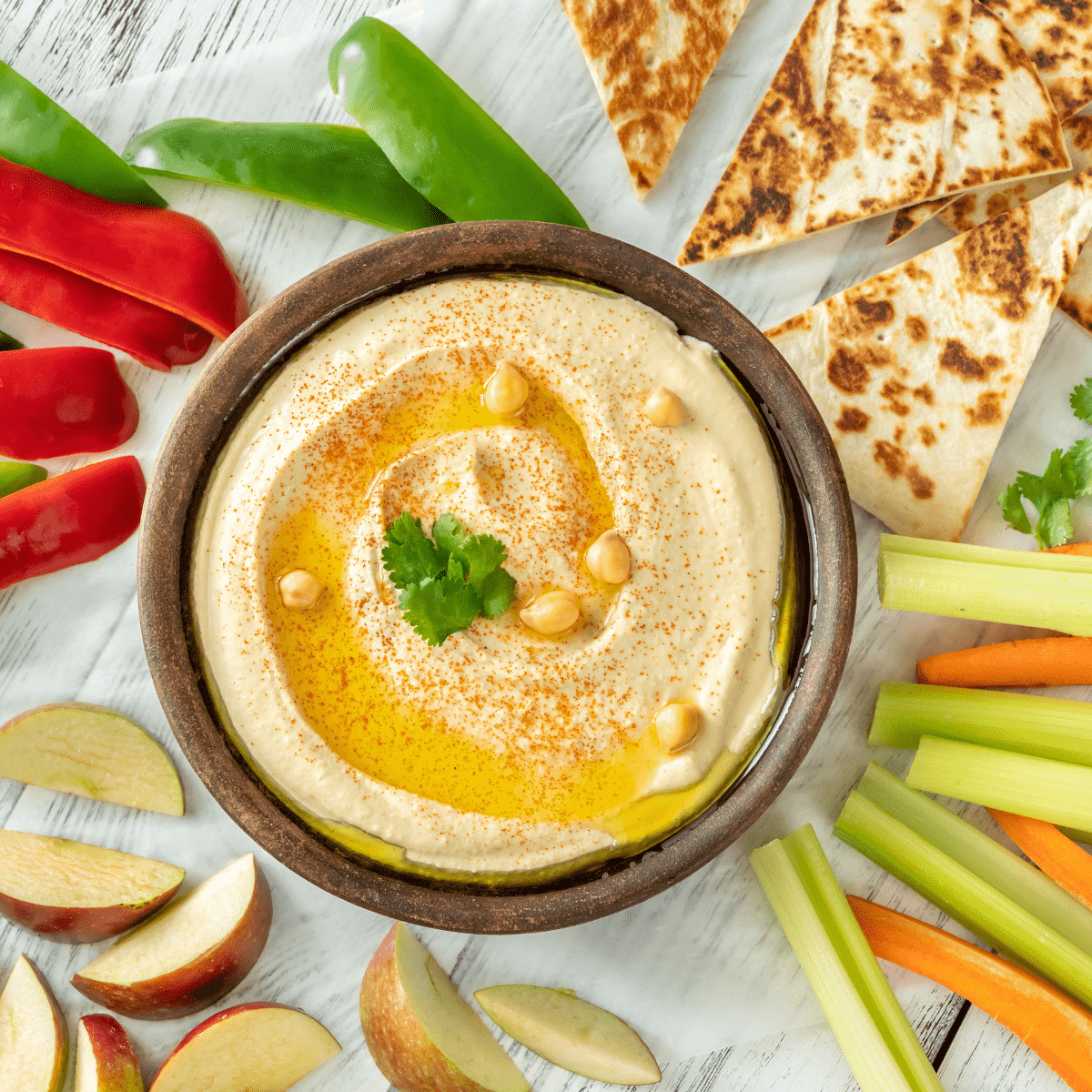 bowl of hummus with sliced peppers, celery, carrots, pita, and apples on white wood board