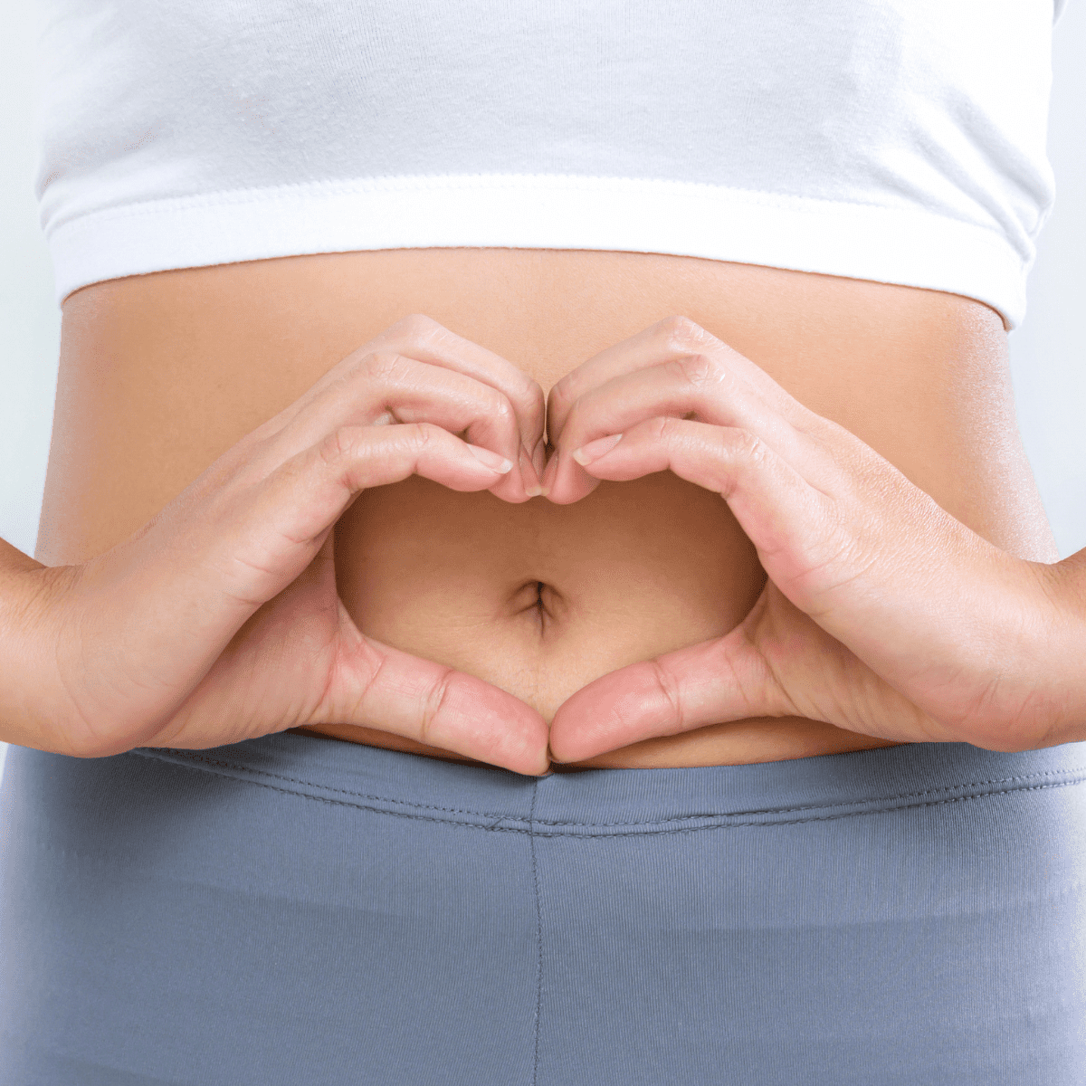 Woman in a white crop top with her hands around her belly button in the shape of a heart. 