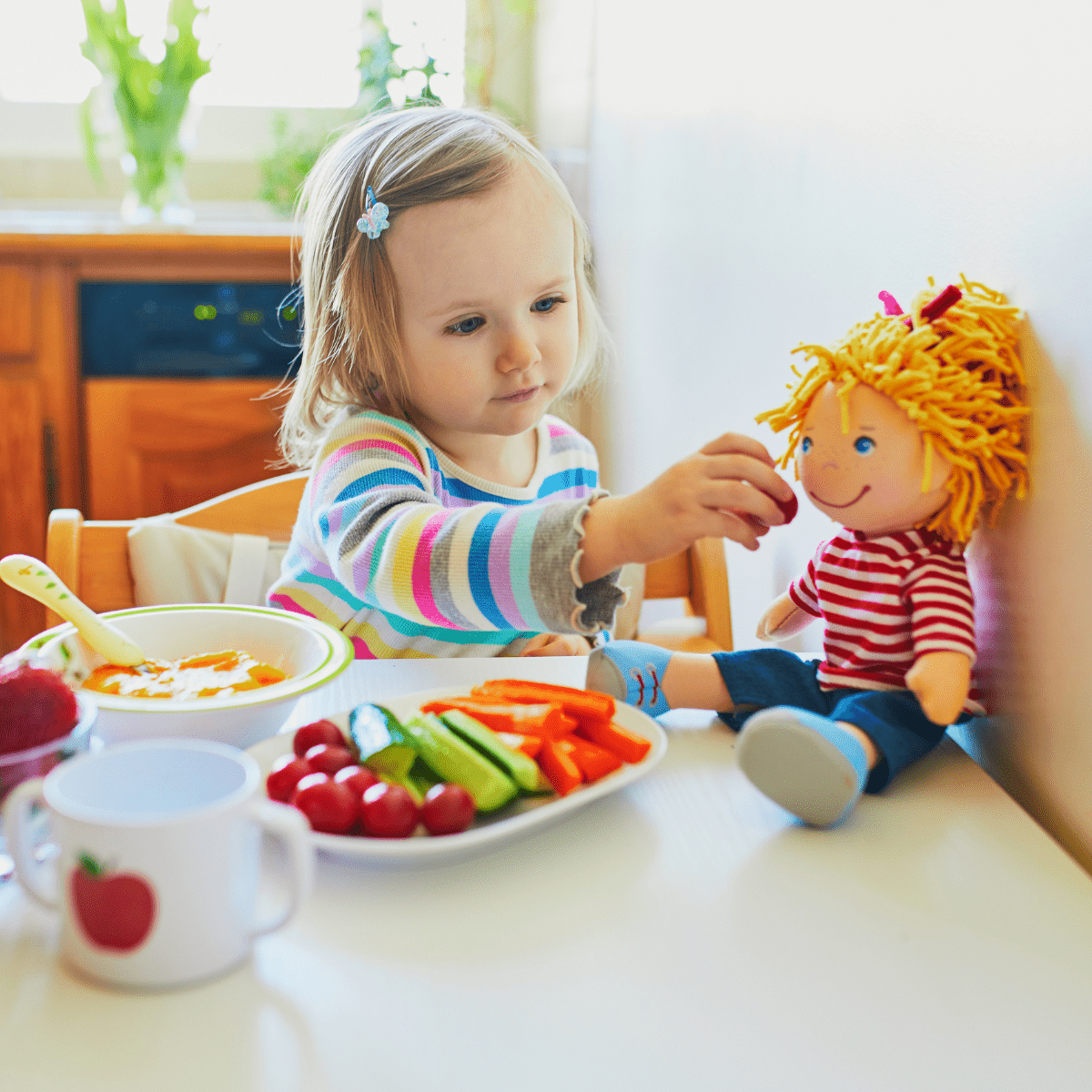 Toddler girl sitting at white table feeding her doll from a plate of vegetable snacks - carrots, cucumbers and tomatoes. 