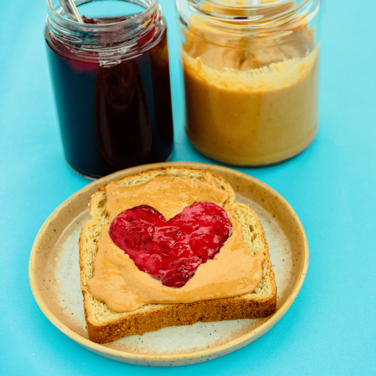 Blue background with glass jars of jelly and peanut butter behind a plate with a piece of bread covered in peanut butter with a jelly heart.