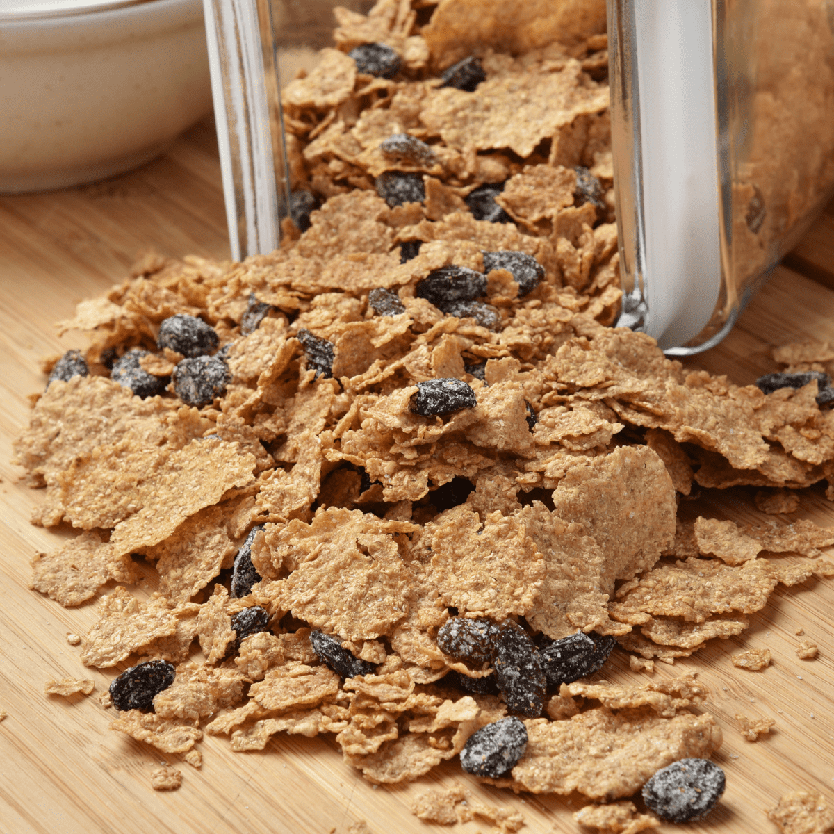 Clear container of bran cereal with raisins spilling out onto wooden countertop. 