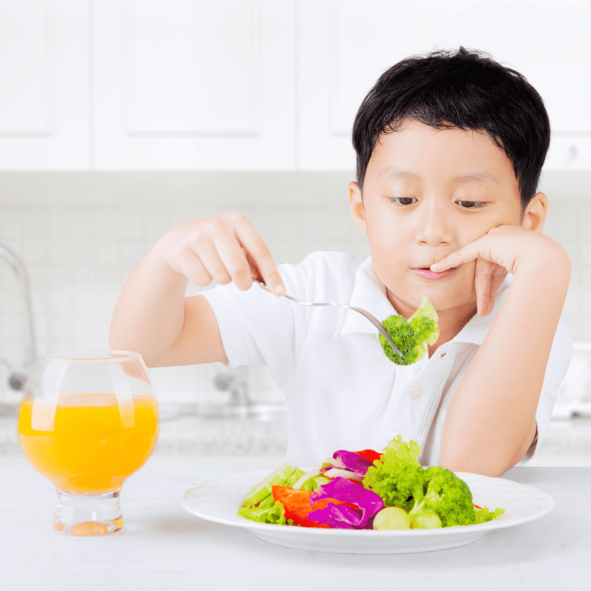 A boy in a white kitchen eating a colorful plate of vegetables with a clear glass of orange juice.