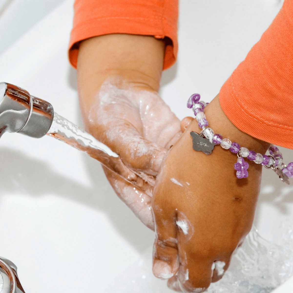 Arms with orange sleeves and a purple and white bracelet washing hands under a running faucet