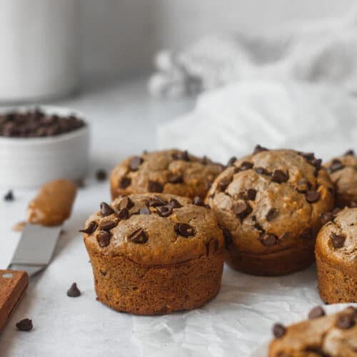 muffins on white tabletop with bowl of chocolate chips and spatula with nut butter