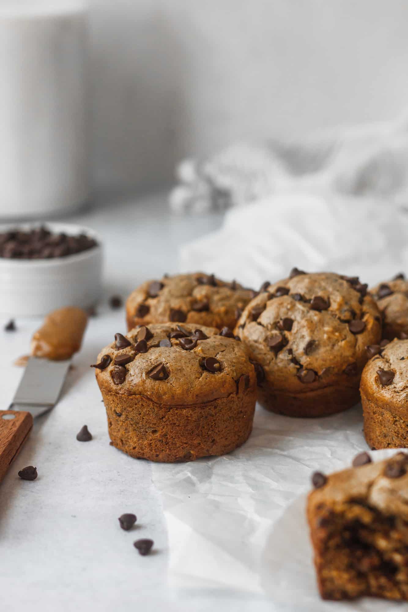 muffins on white tabletop with bowl of chocolate chips and spatula with nut butter