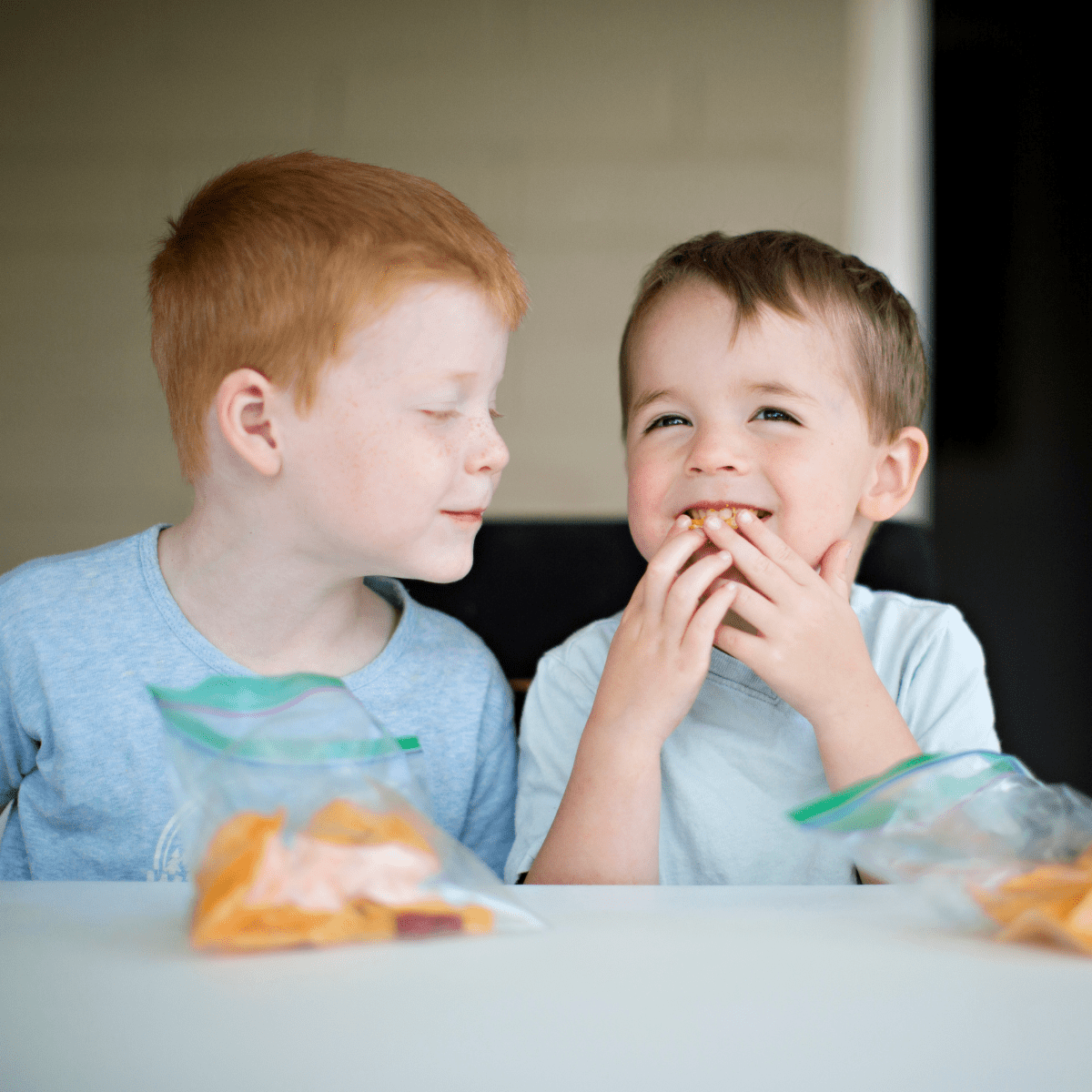 Two boys sitting at a table eating an orange snack out of plastic bags.