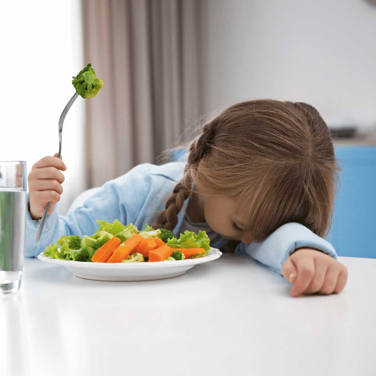 Young girl with head down on table, next to a plate of vegetables, holding a fork in the air with a piece of broccoli on it