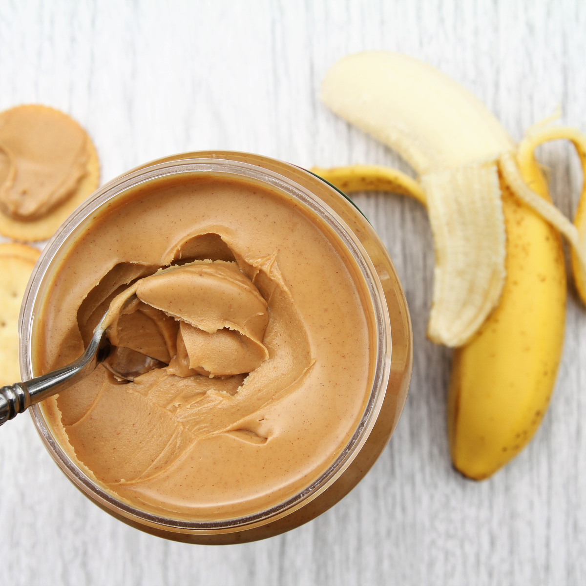 Half-peeled banana on a table next to a jar of nut butter on a tabletop. Round crackers in the background with nut butter spread on one cracker.