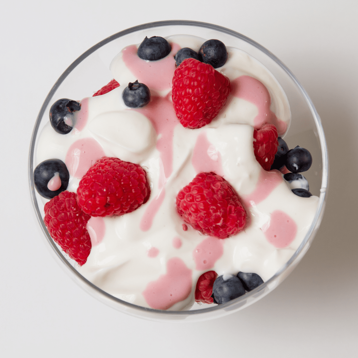 Clear bowl on white table, filled with white yogurt with blueberries, raspberries and pink fruit juice drizzled on top 