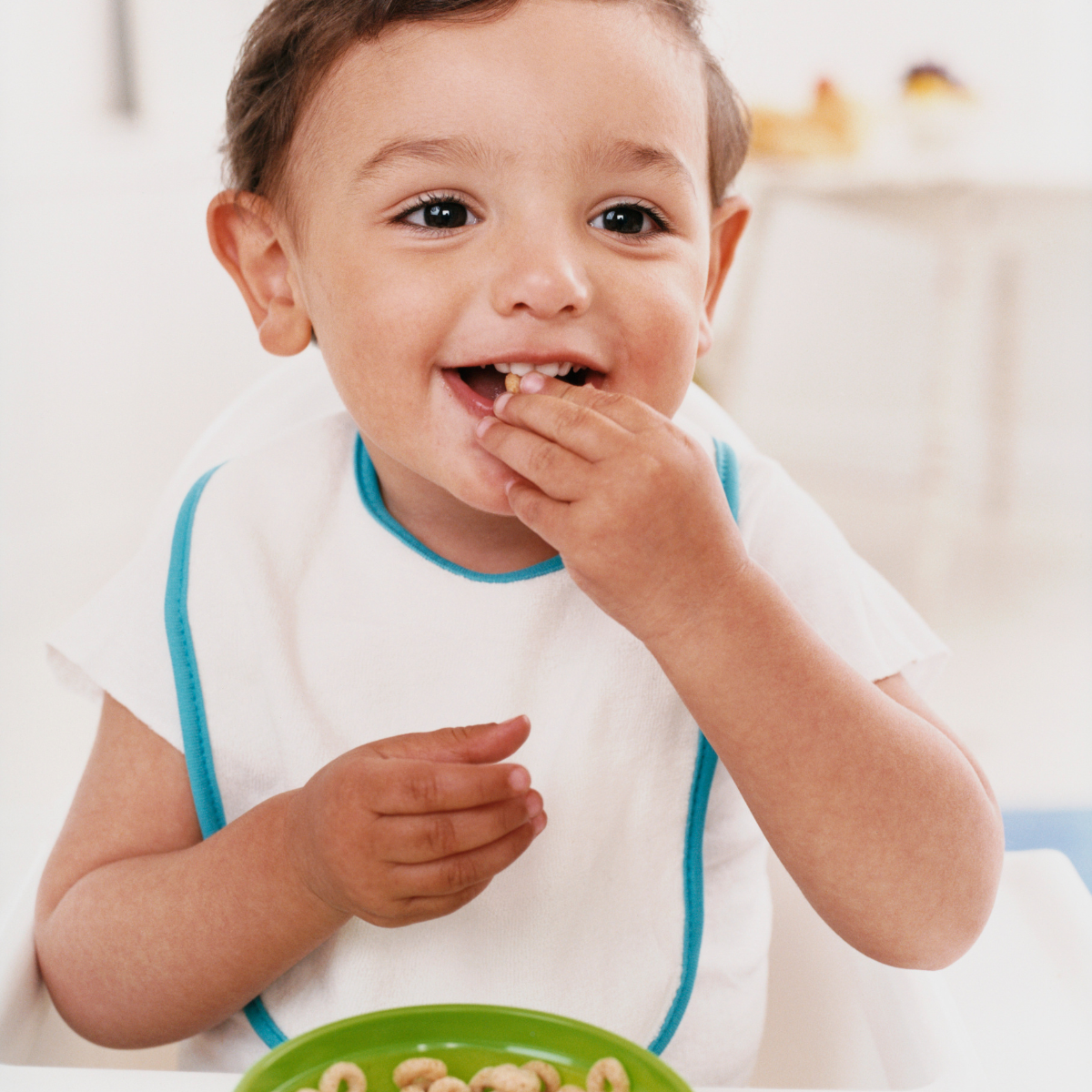 Toddler boy with white shirt, white bib and green plate of cheerios eating with his hands.