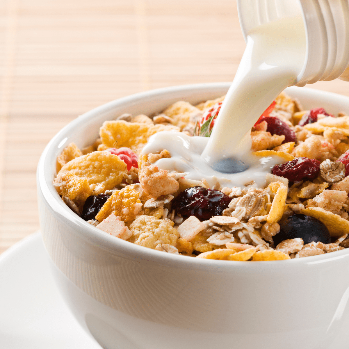 White bowl of cereal with fresh and dried fruit and nuts with milk being poured into bowl. 