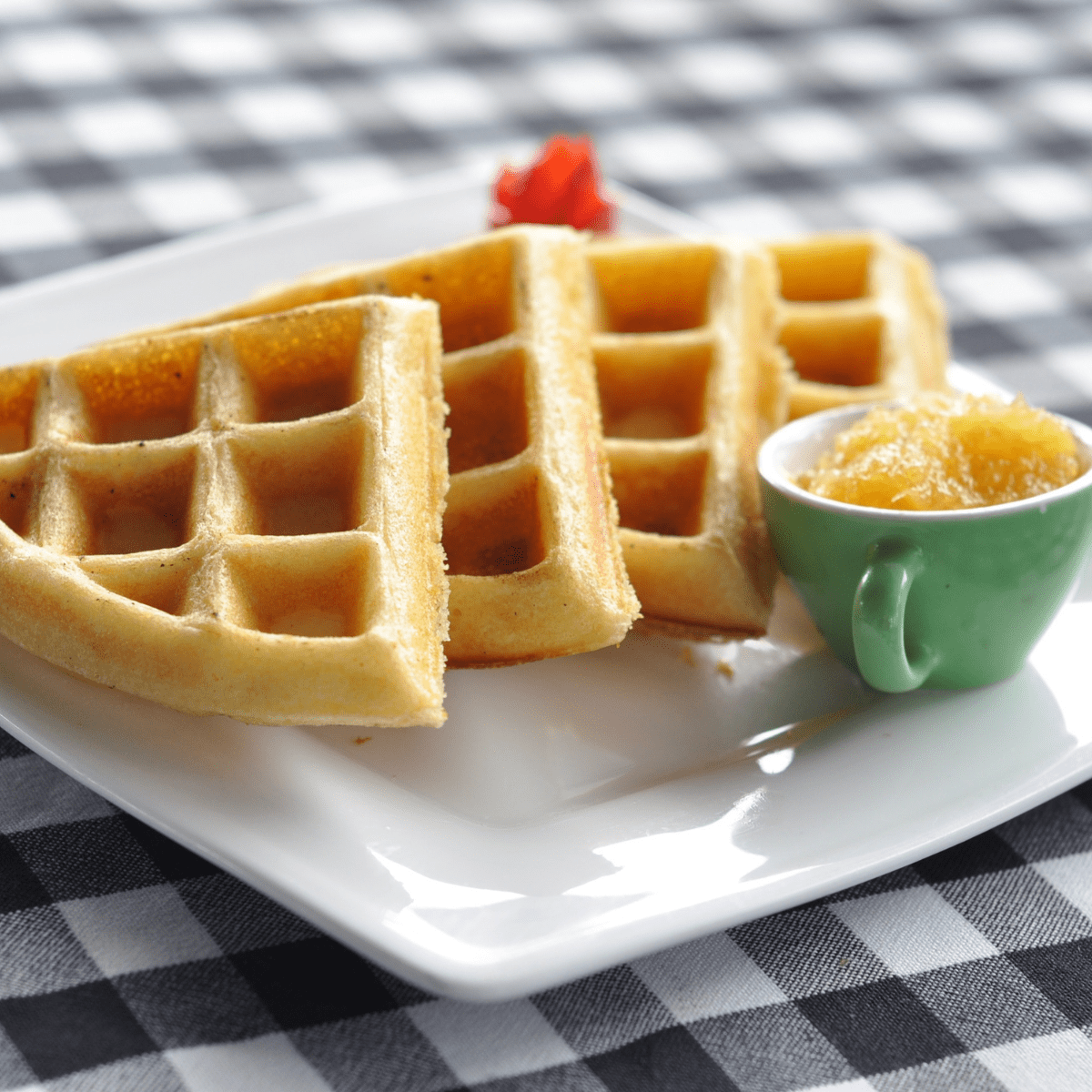 Waffles on a white plate with a small dish of applesauce on the side, served on a white plate on a white and black checkered tablecloth. 