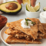 Triangle-cut baked quesadillas on white plate with a hand squeezing a lime