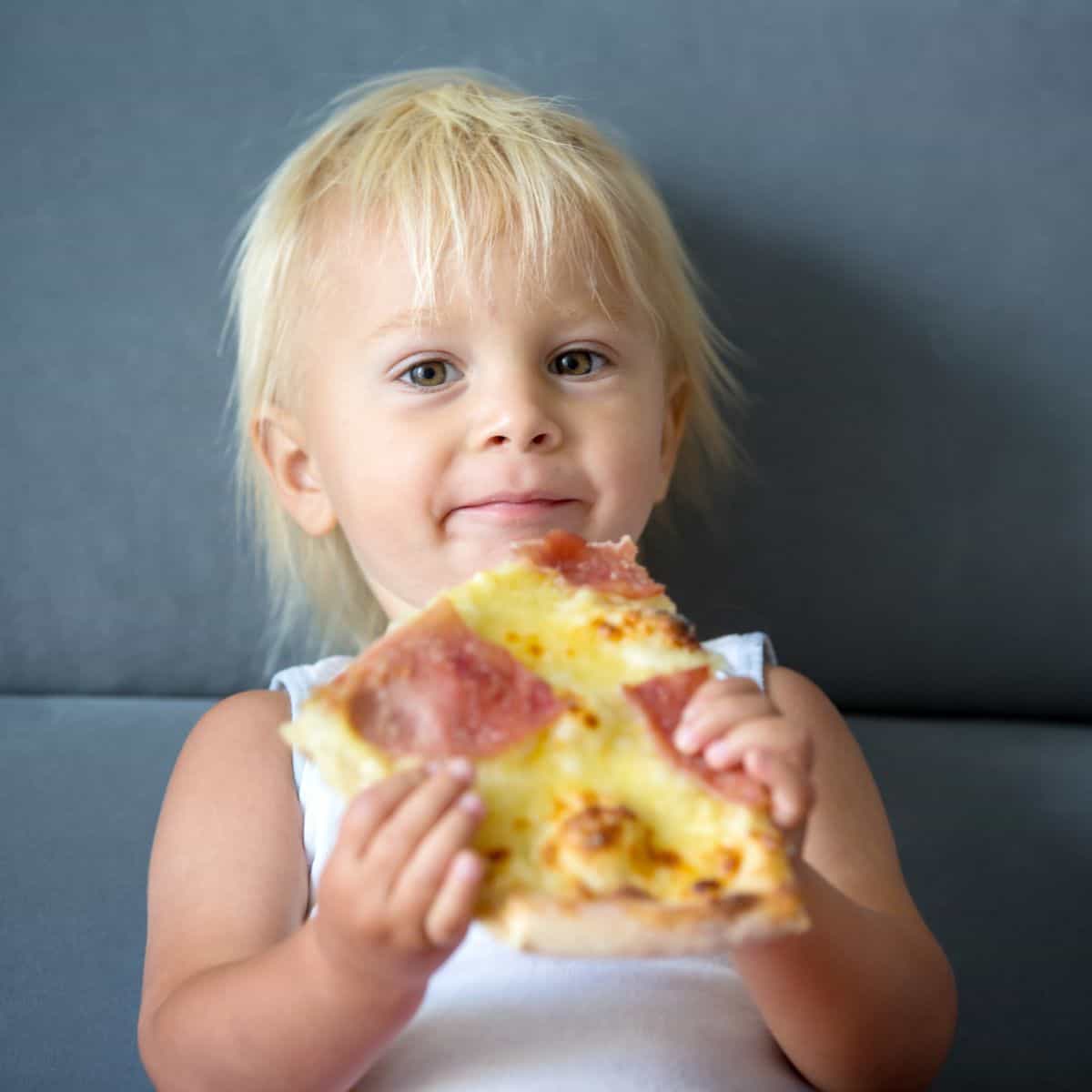 Toddler sitting against a solid background with a slice of pizza in their hands