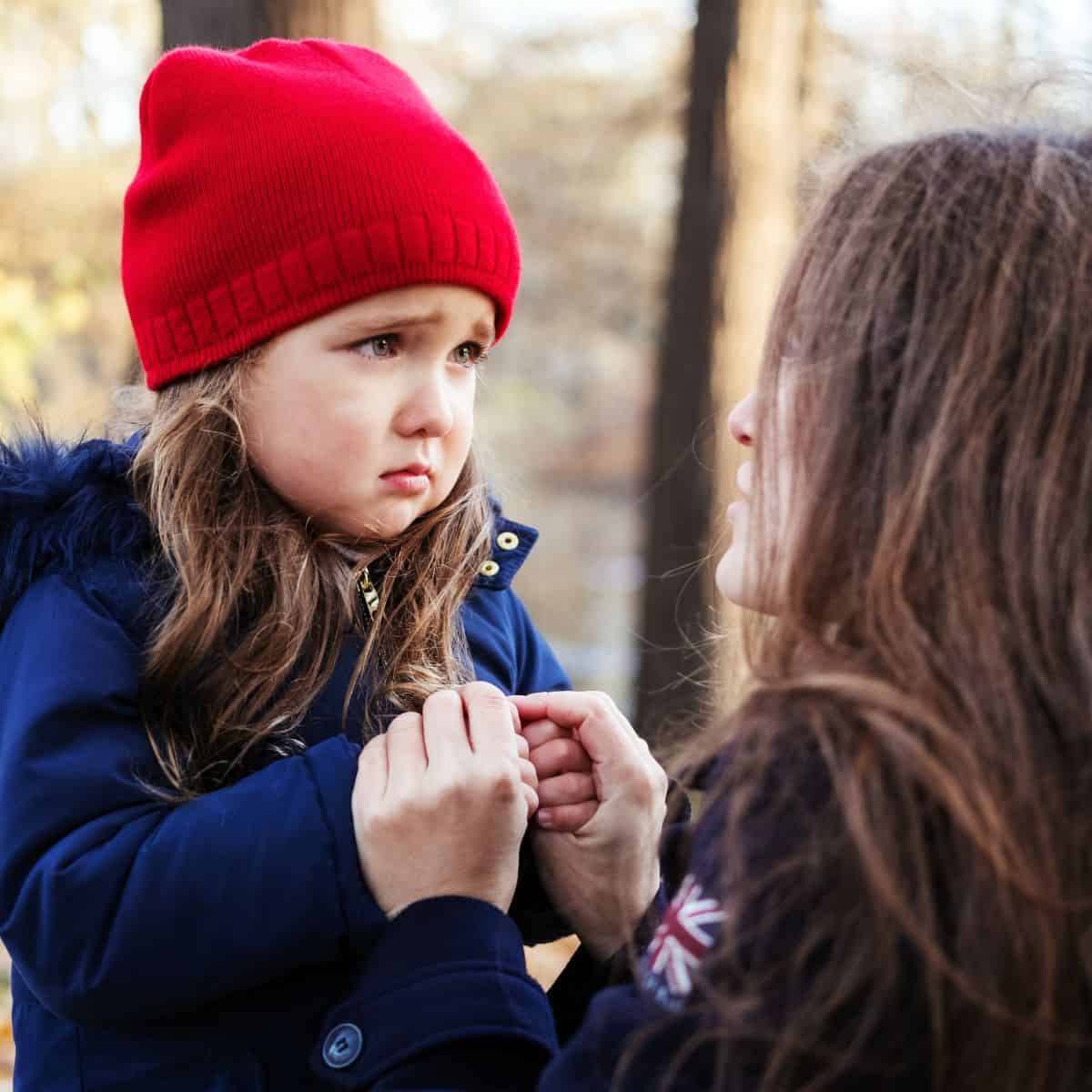 preschool child in red hat and blue winter coat looking sad with mom holding her hands