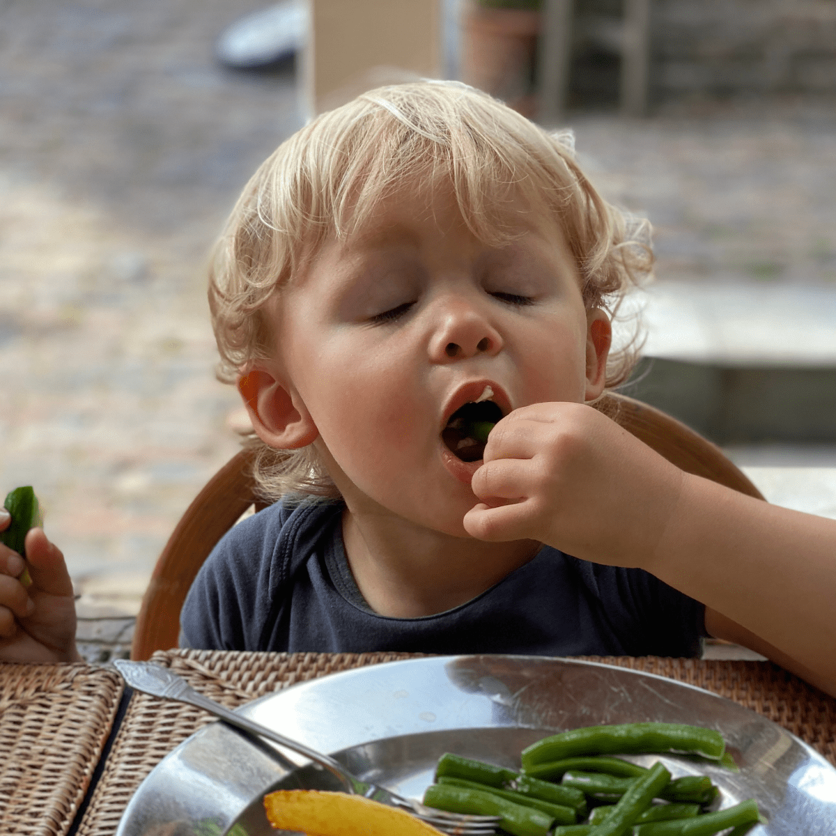 Toddler boy eating veggies with his eyes closed and mouth open to take a bite