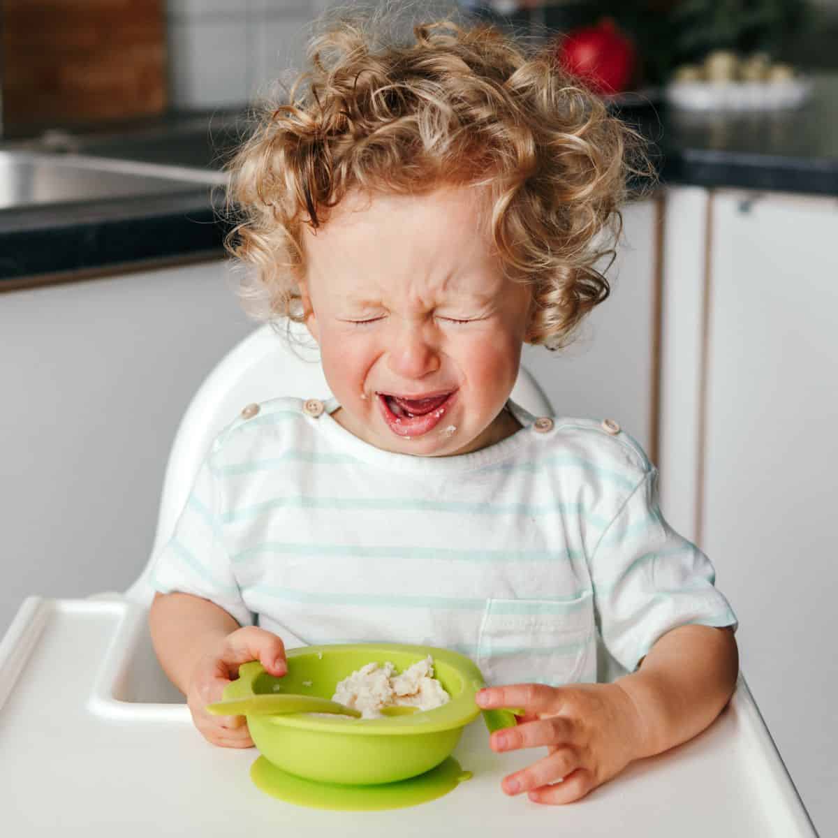 Toddler with eyes closed, crying, while sitting in a high chair with a bowl and spoon sitting in front of them.