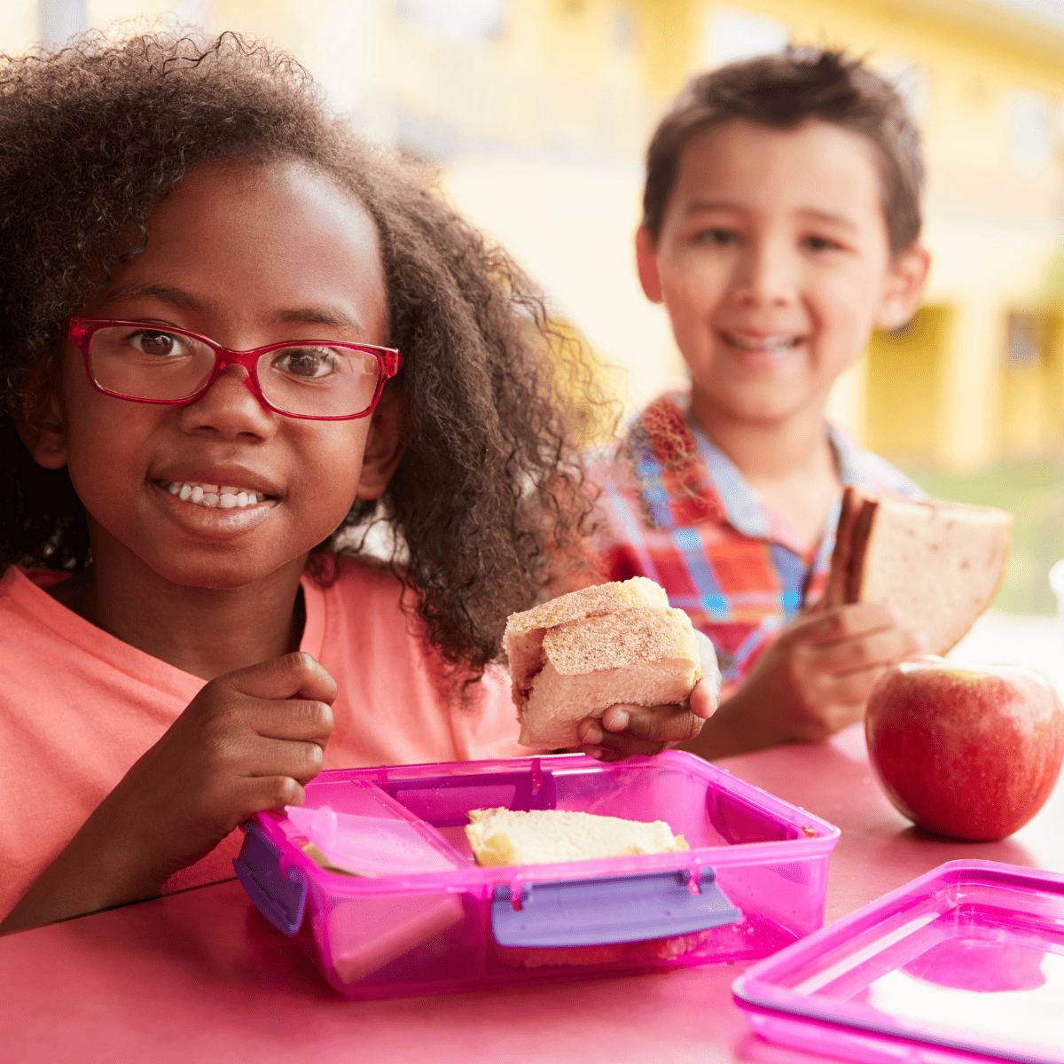 two children eating lunch together, pink lunchbox on the table